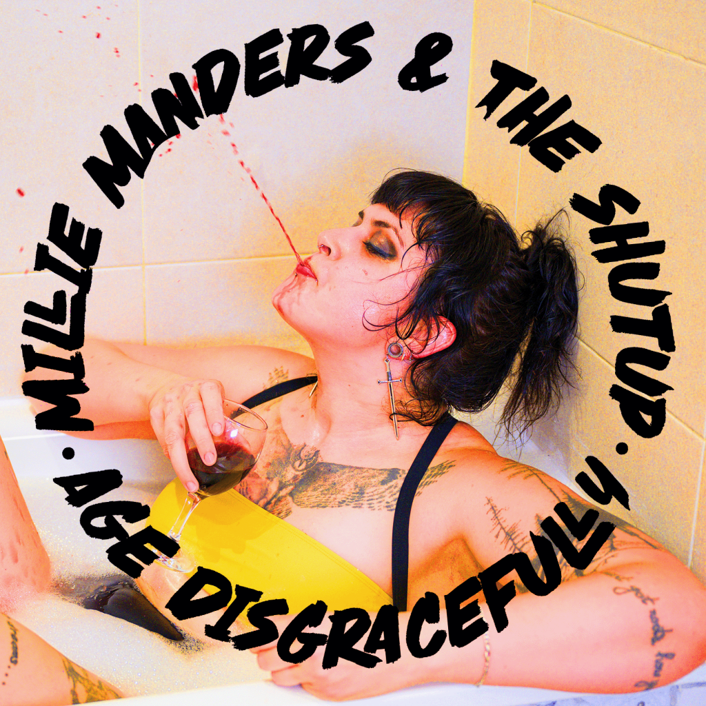 A woman with dark hair and tattoos lounging in a bathtub holding a glass of red liquid, with a playful expression. Text overlay reads 'Millie Manders & The Shut Up - Age Disgracefully'.