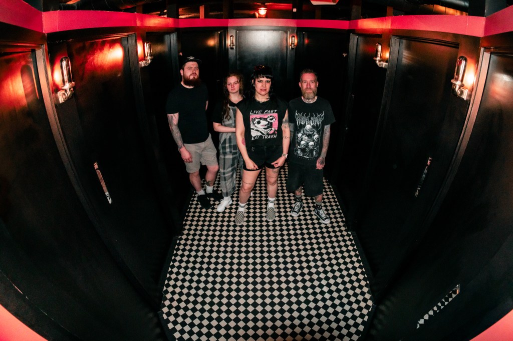 A group of four individuals standing in a modern restroom with black walls and a checkered floor, illuminated by dim, vintage-style lights.